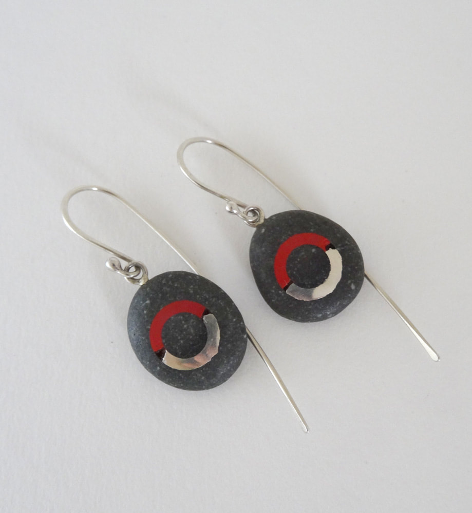 These Earrings are made from Sterling Silver and Greywacke Pebbles. Maike collected the pebbles on Birdlings Flat beach on the South Island of New Zealand. She then carved a circle into the stones and filled it with molten silver and later with low firing red enamel.